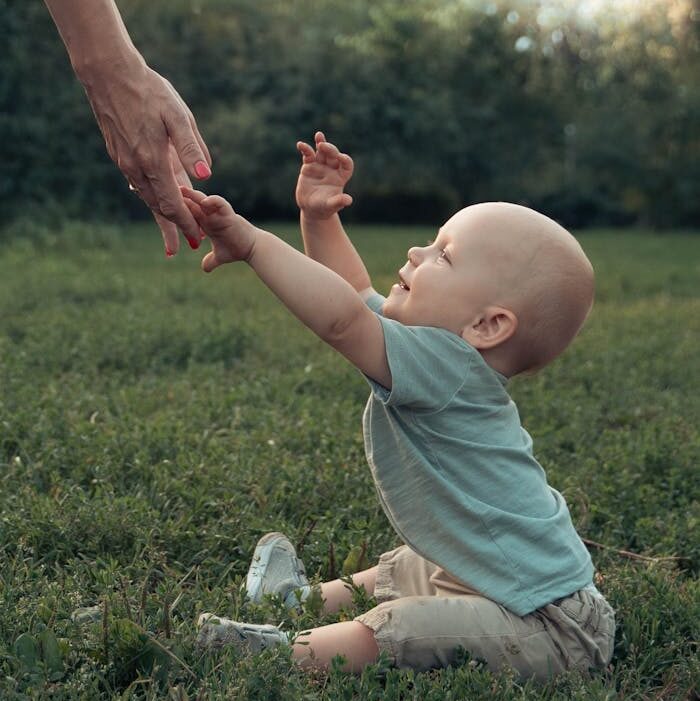 A cute toddler reaches for a parents hand while sitting on grass in a Moscow park.
